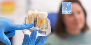 Close-up of a dental model showing implants and artificial teeth held by a gloved dentist, with a patient in the background at Dentistry on Sinclair, highlighting the benefits of dental implants